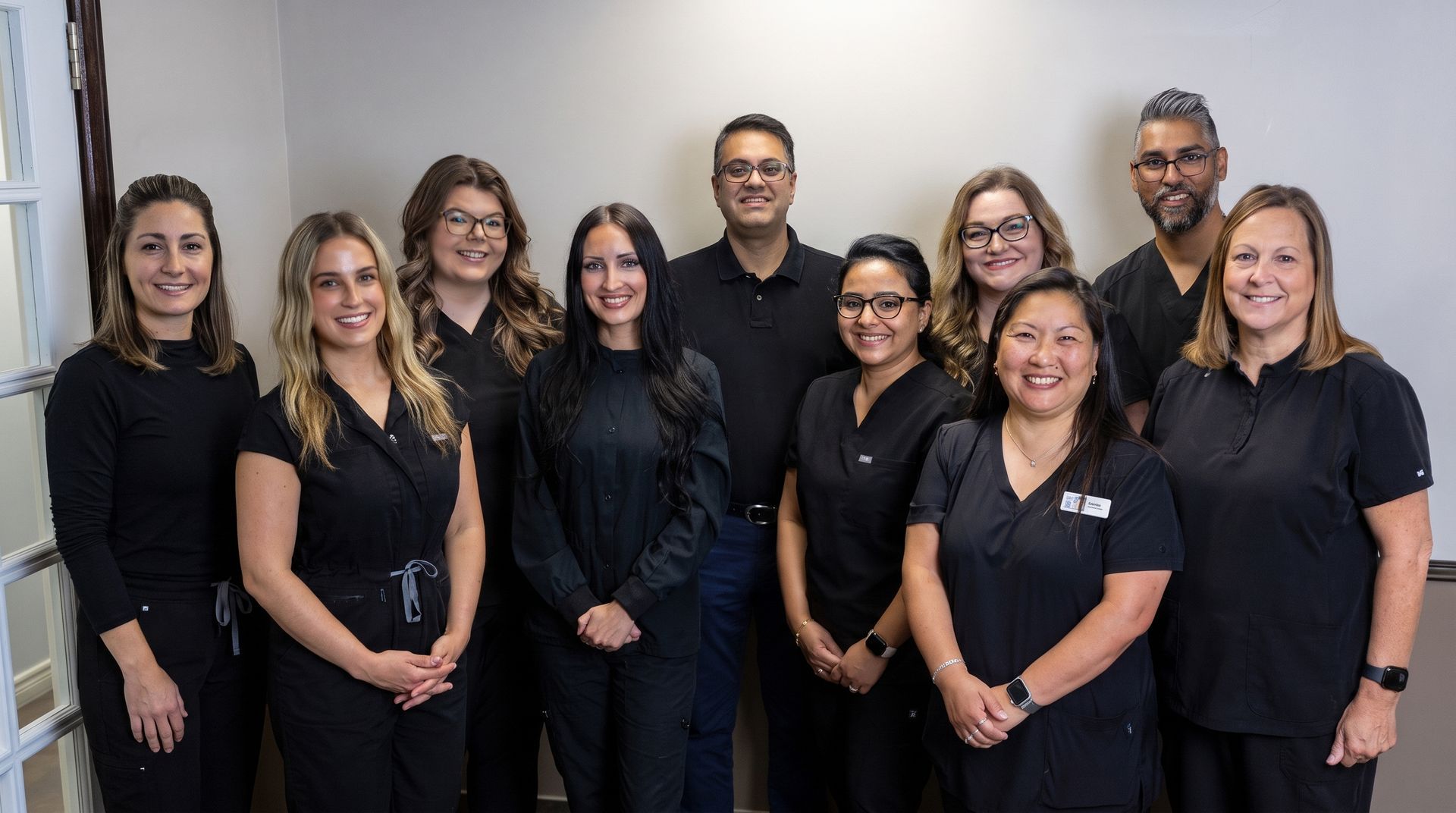 Group of eight people in black scrubs, smiling, posing in an office setting with a gray wall.