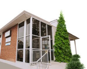 Brick building with large windows and open door; evergreen tree to the right, sign on the left.