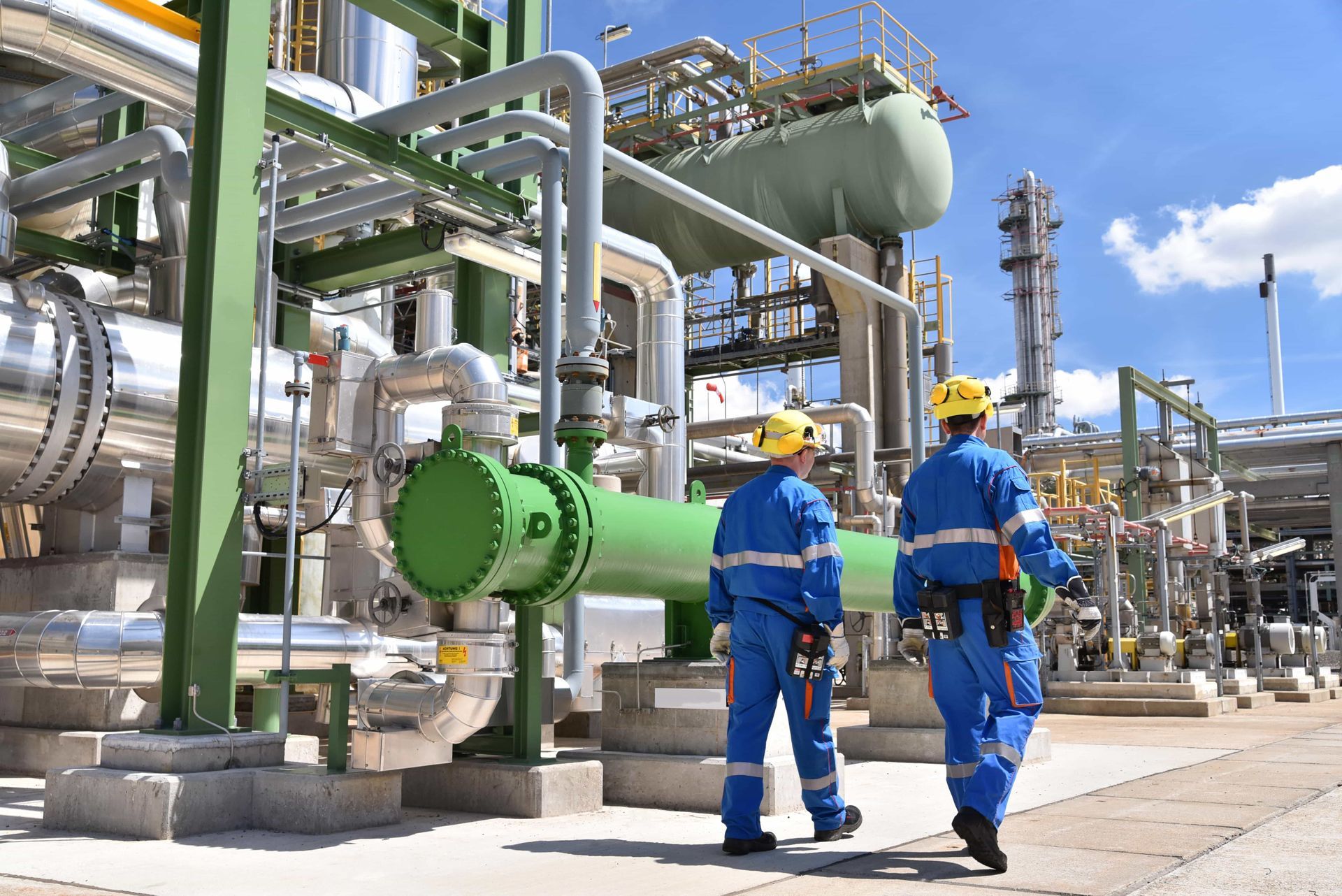 Two men are walking in front of a large industrial plant.