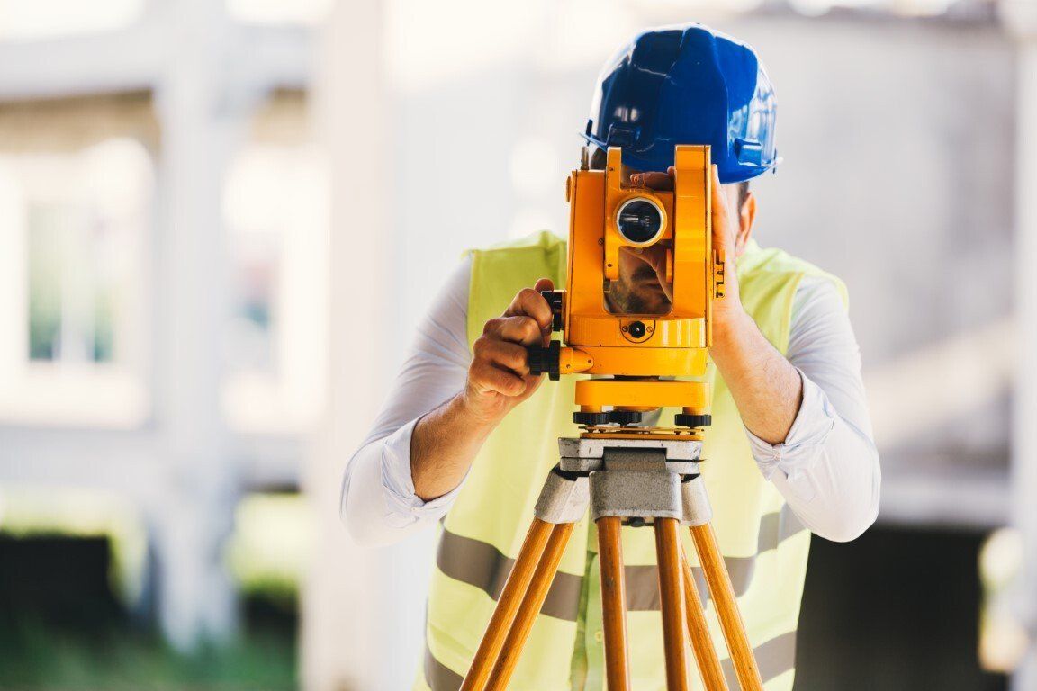 A man is using a theodolite on a tripod on a construction site.