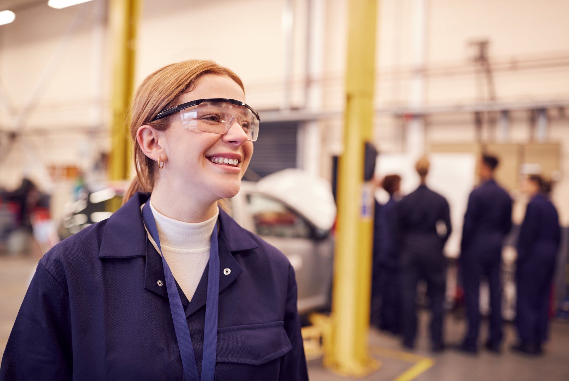 A woman wearing safety glasses is smiling in a garage.