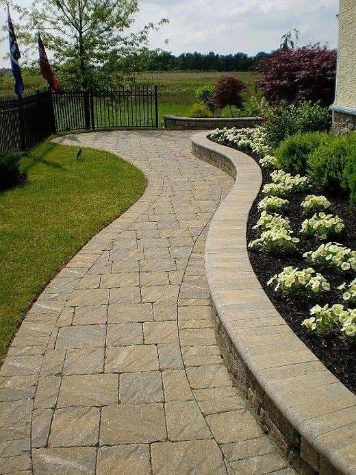 Stone path curves through a landscaped yard with flower beds and green lawn, leading to a black gate.