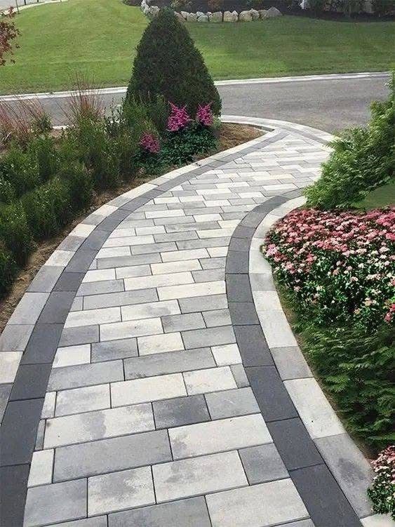 Curving paved walkway bordered by greenery, leading to a landscaped yard with flowers and a tree.