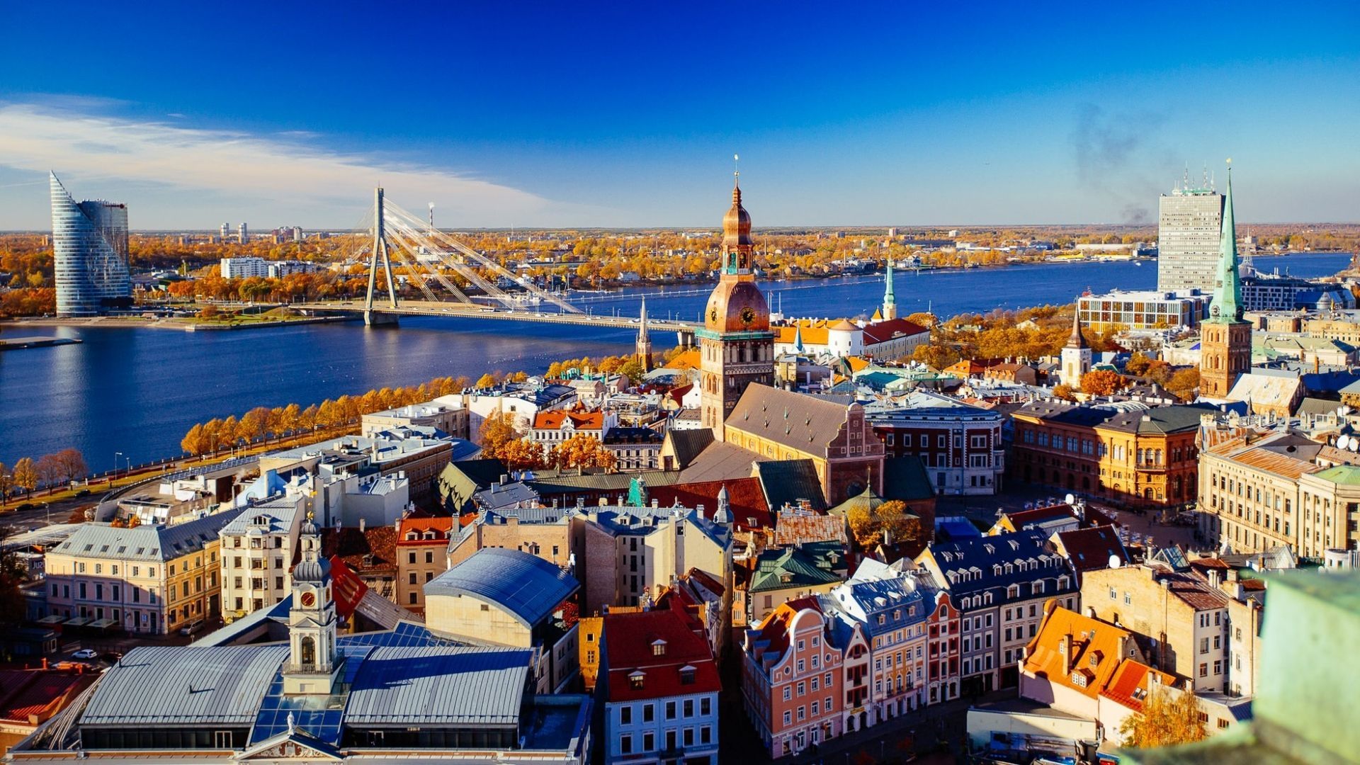 Panoramic waterfront cityscape with colorful rooftops, church towers, and a bridge over water under a blue sky