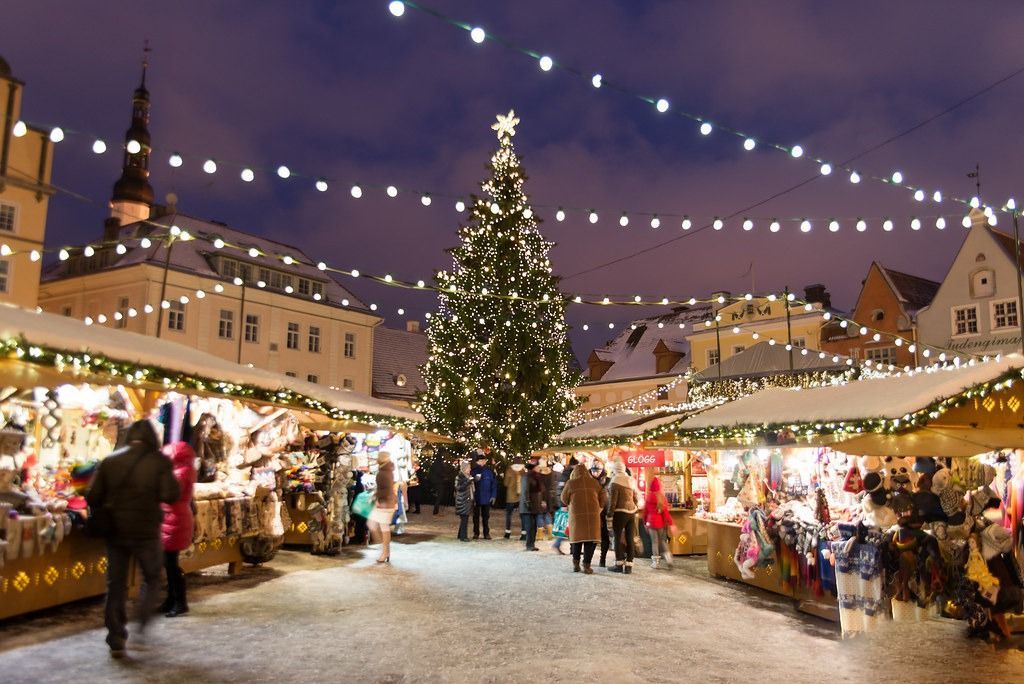 Twinkling Christmas market at night with decorated stalls, a lit tree, and people walking along snowy path