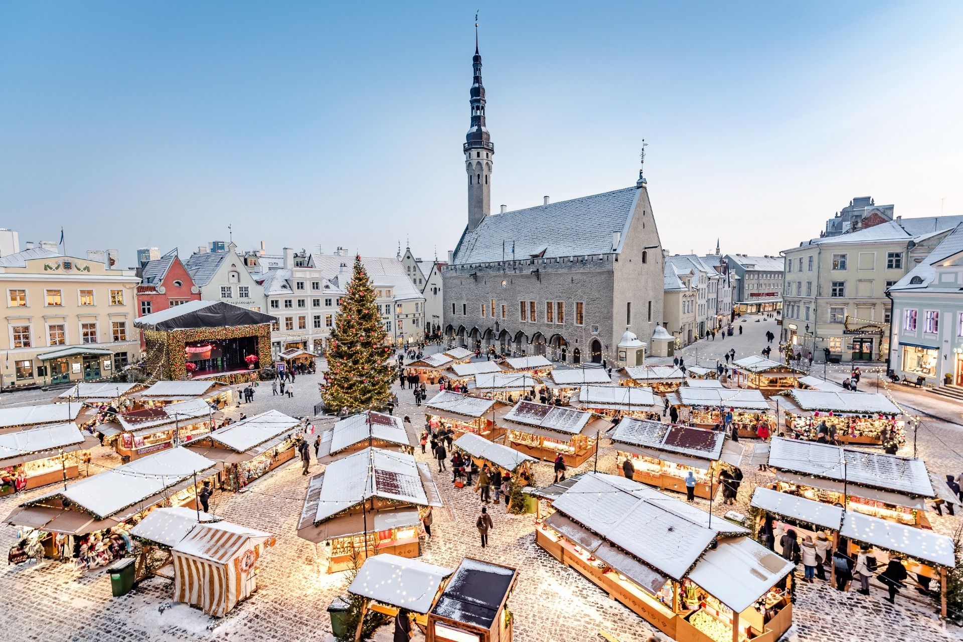 Snowy Christmas market with lit wooden stalls and a tall church tower in a historic town square