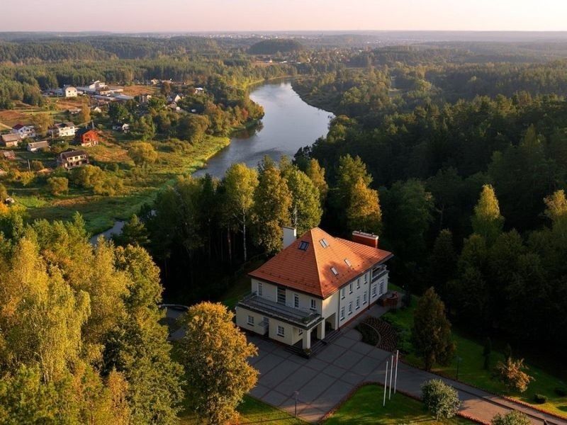 Aerial view of a red-roofed building by a river, surrounded by dense green forest at sunset
