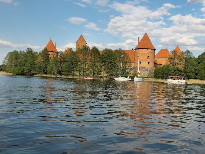 Red-roofed castle by a lake, framed by trees under a partly cloudy sky.