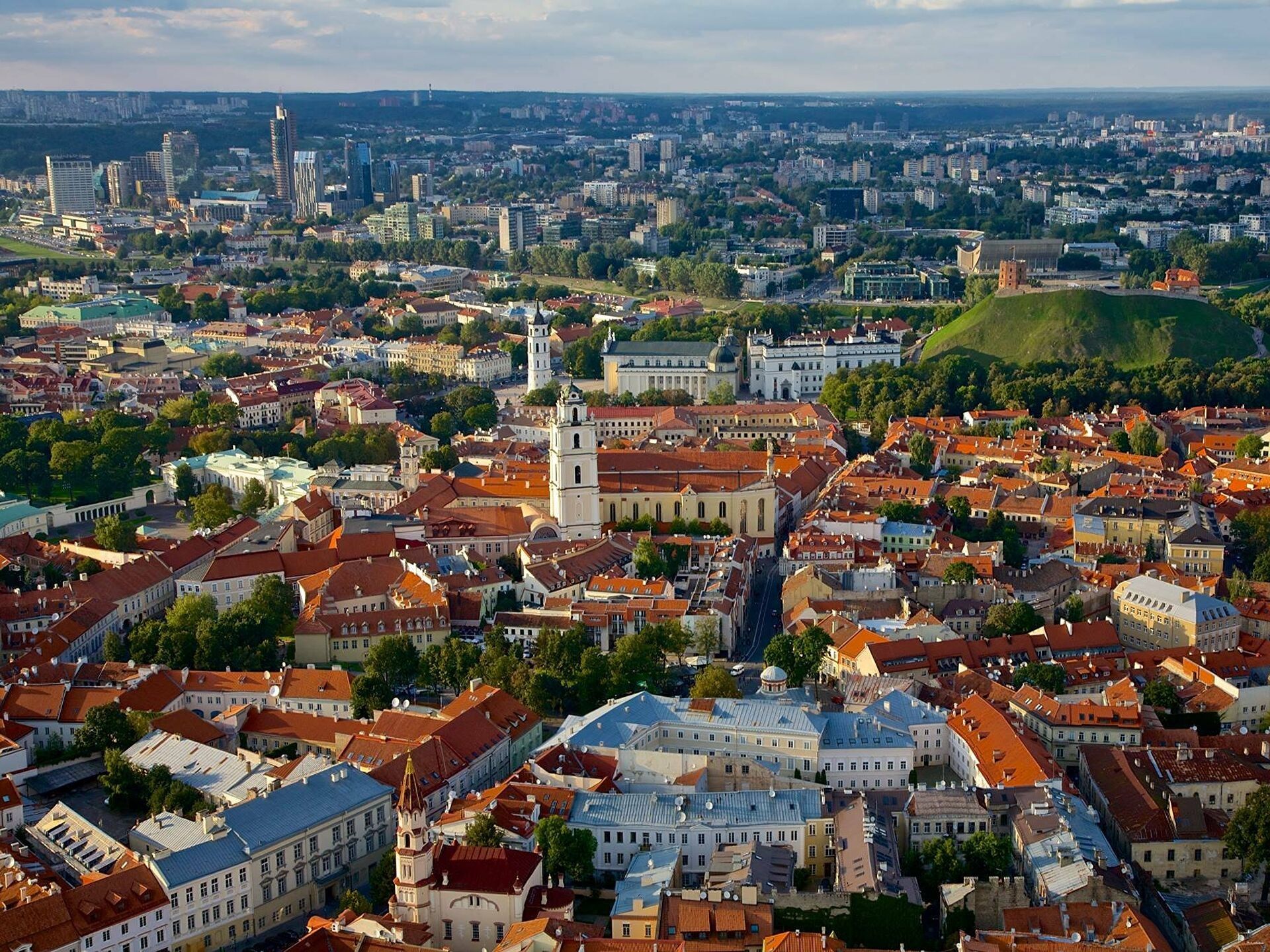 Aerial view of a historic city with red rooftops, a white clock tower, and a distant skyline under clear skies