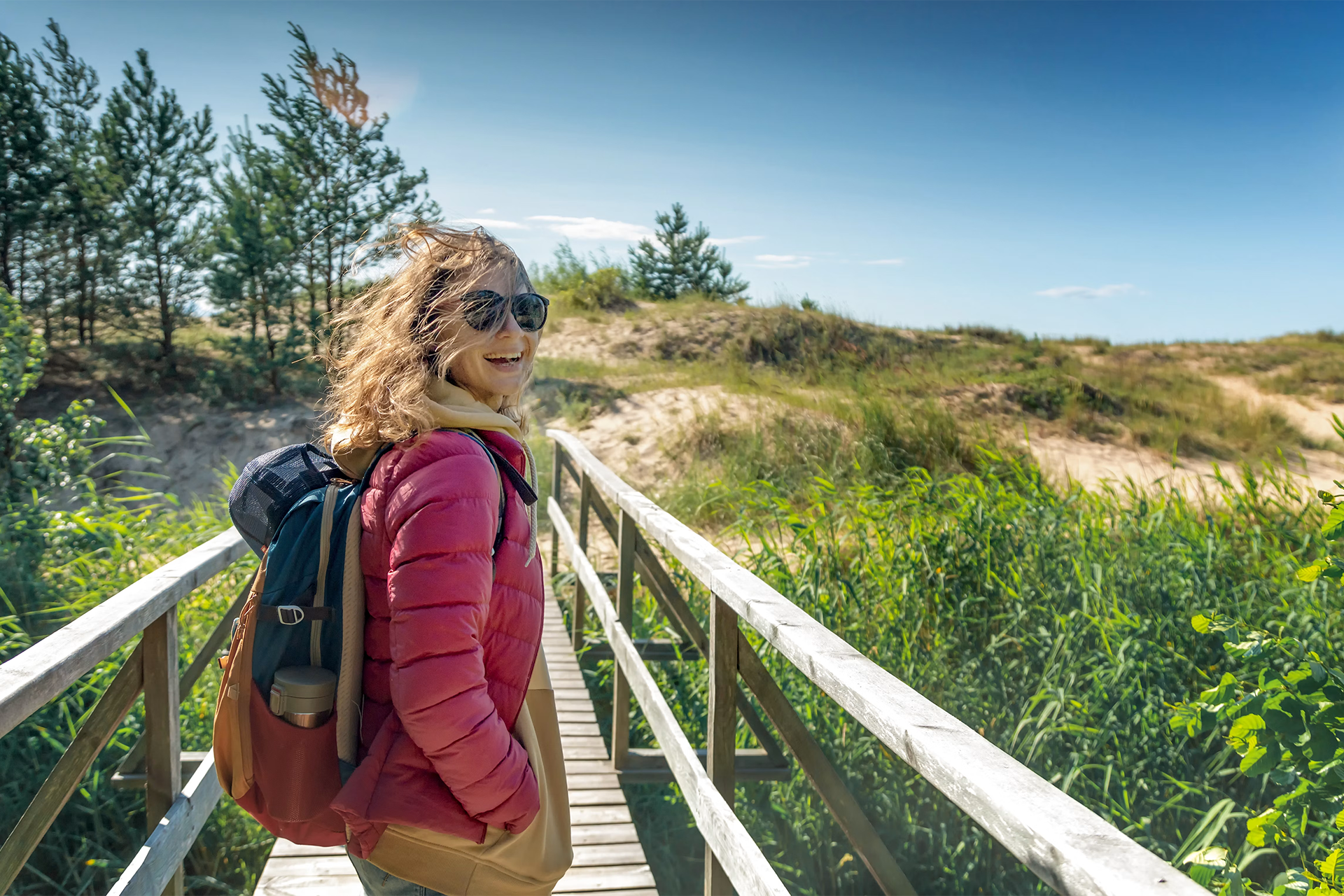 Woman with backpack on a wooden boardwalk through sandy dunes and tall grass, smiling in the sunlight