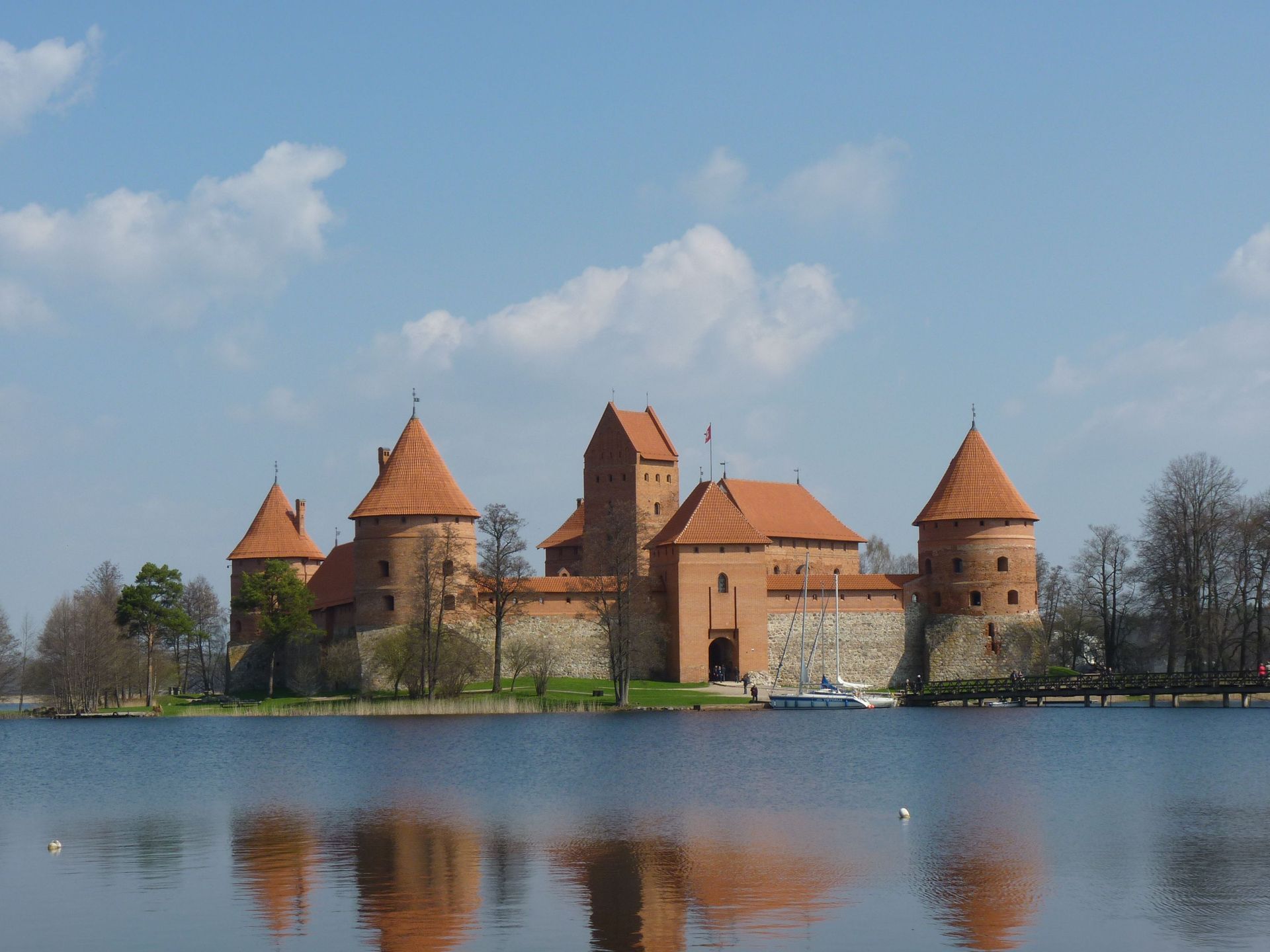 Stone castle with red roofs beside a calm lake, reflected in the water under a blue sky