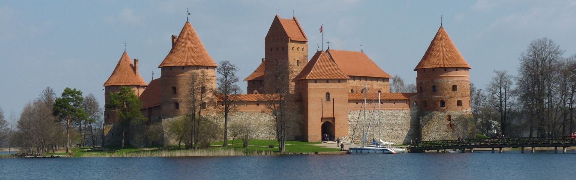 Brick castle with red-roofed towers beside a calm river, with trees and a small boat nearby