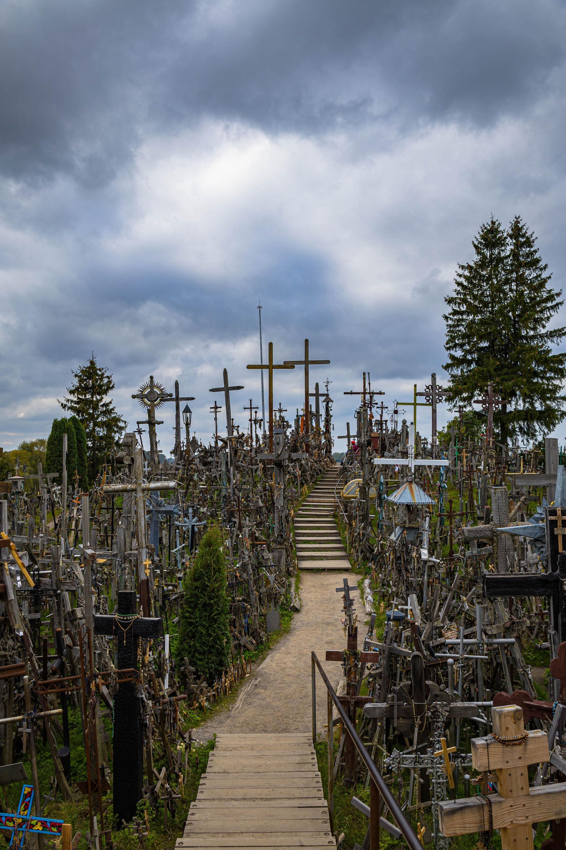 Crosses and memorials along a narrow path under a cloudy sky in a forested hillside cemetery