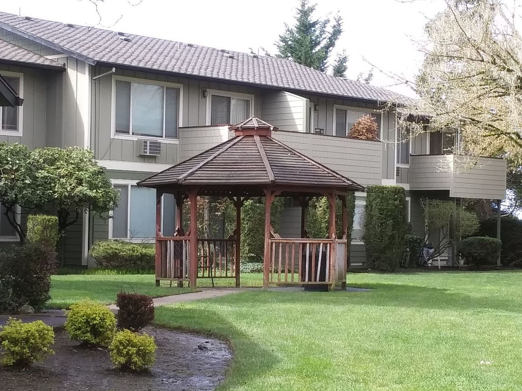 Apartment courtyard with a wooden gazebo, green lawn, and shrubbery in front of two-story buildings