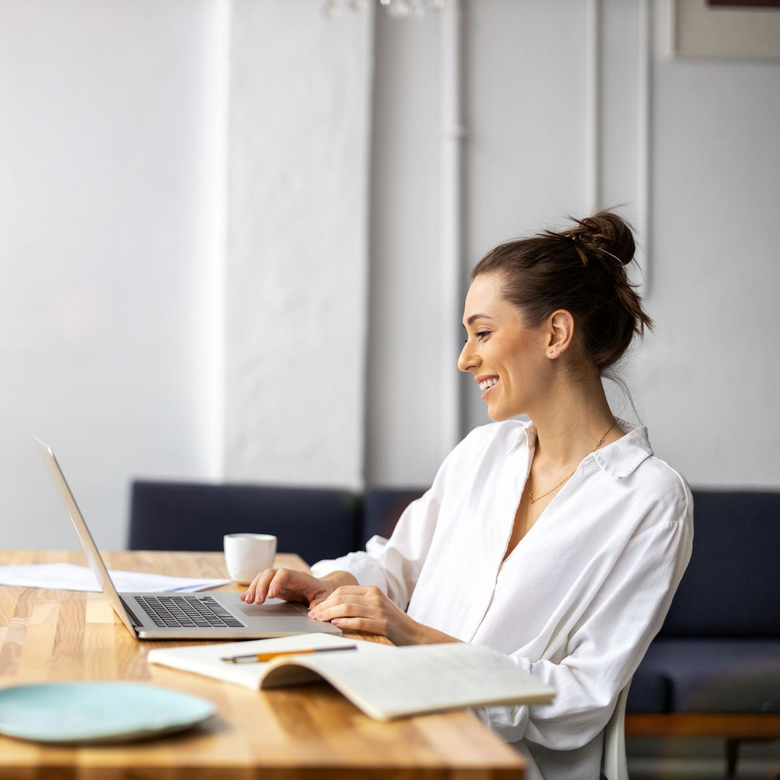 Woman working on a laptop at a wooden table with a notebook and coffee in a bright office.