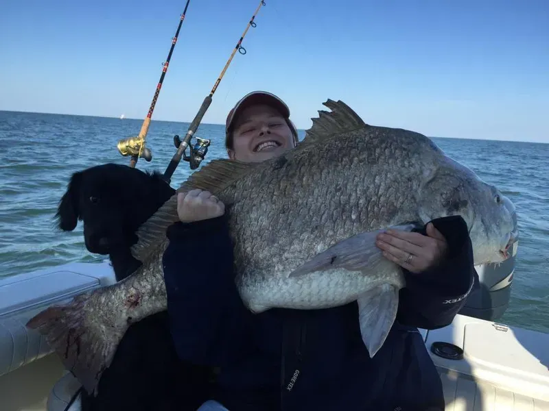 Woman smiling, holding large gray fish on a boat. Black dog sits next to her; fishing poles in background.