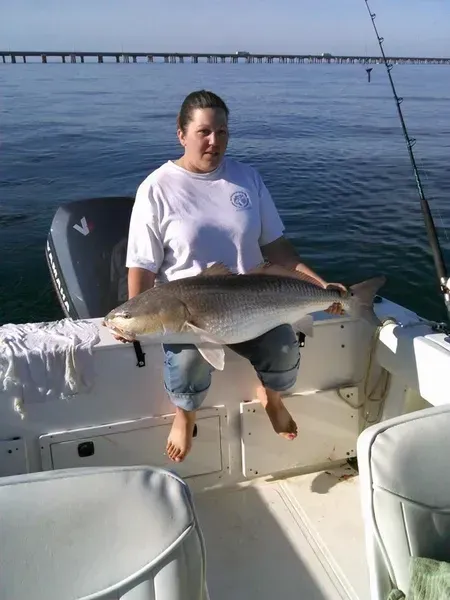 Woman on a boat holding a large redfish; sunny day, water, bridge in background.