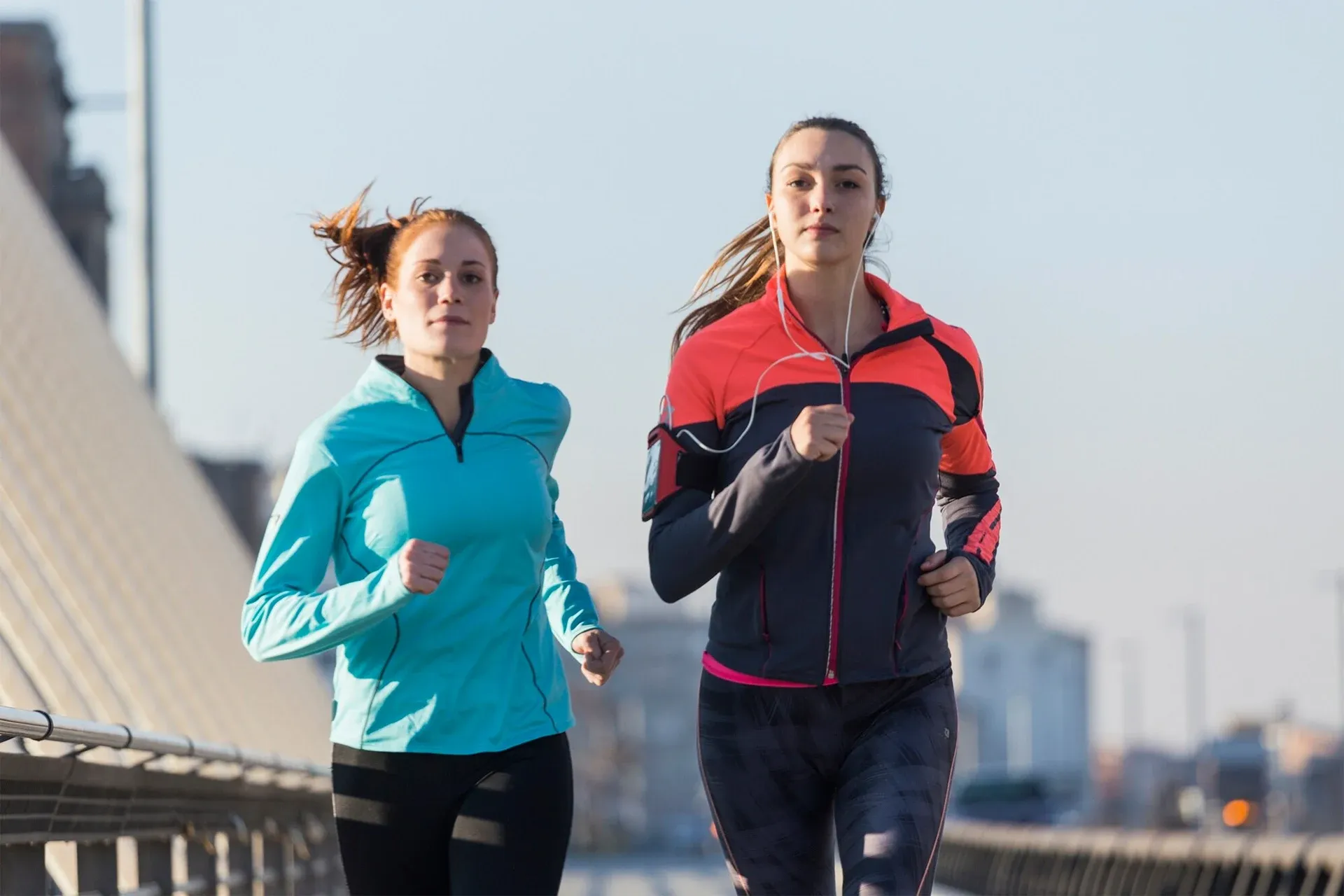 Two runners jogging outdoors on a bridge, one in teal and one in red and navy jackets.