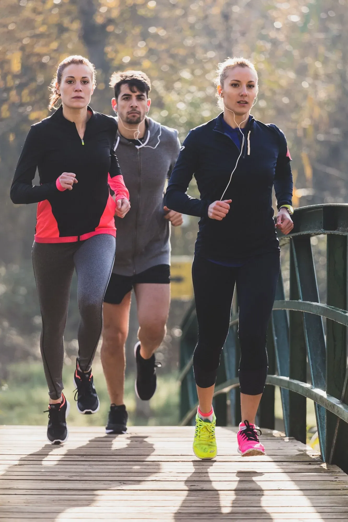 Three runners jog across a wooden bridge in athletic wear, with autumn trees blurred in the background.