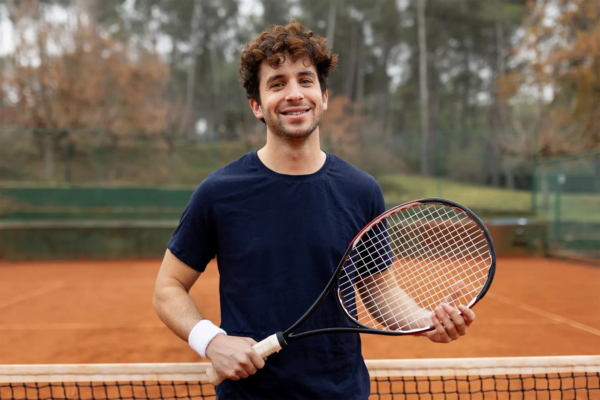 Tennis player smiling on a clay court, holding a racket near the net with a tennis ball in hand.