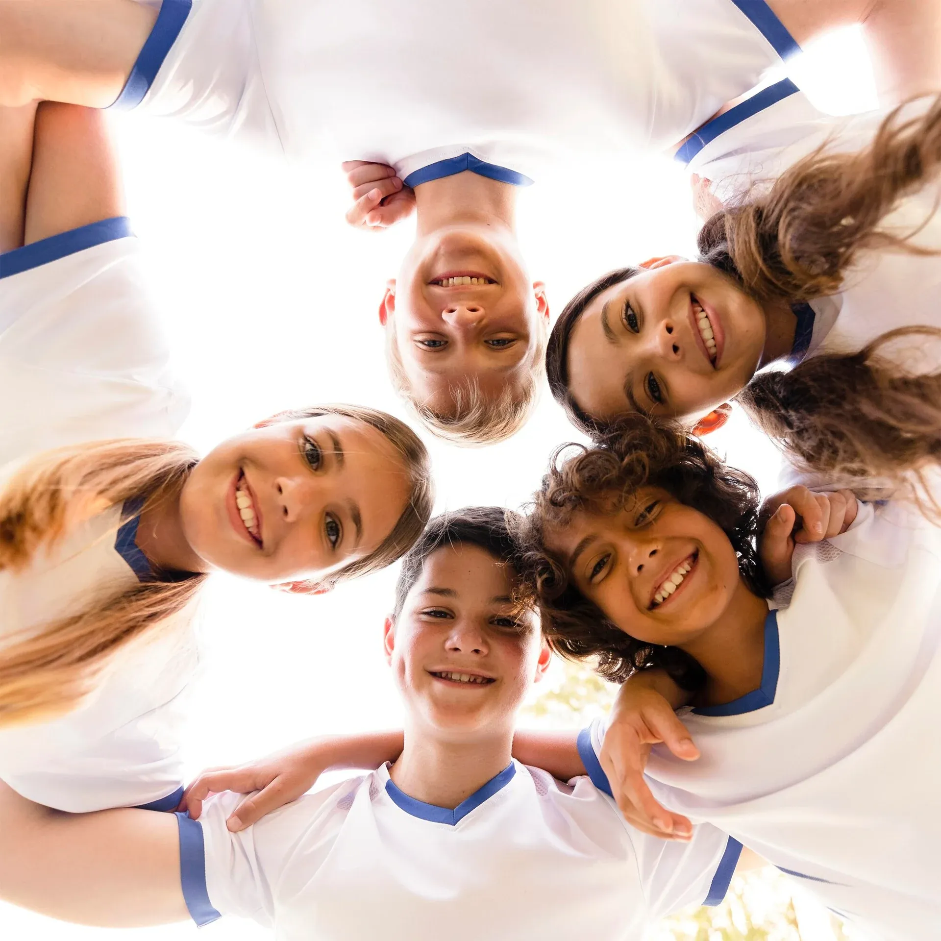 Five smiling children in white-and-blue shirts huddle in a circle, looking down at the camera.