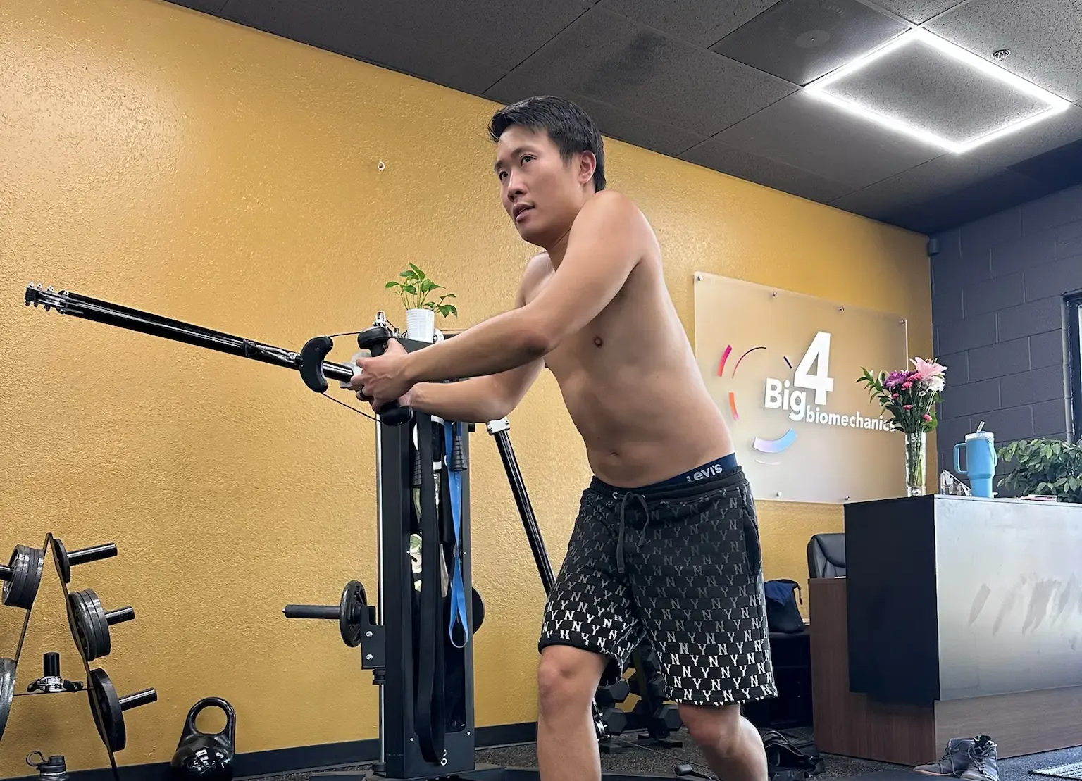 Man posing shirtless with a sword-like prop in a gym, wearing black patterned shorts.