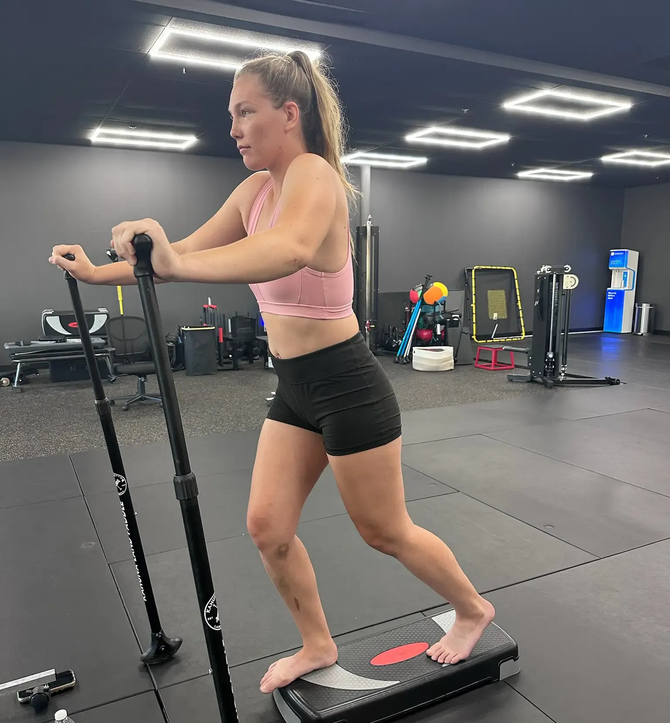 Woman exercising on a stair-stepper in a gym, wearing a pink top and black shorts.