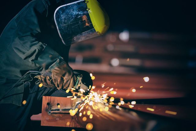 Un saldatore con indumenti protettivi produce scintille metalliche in un'officina poco illuminata.