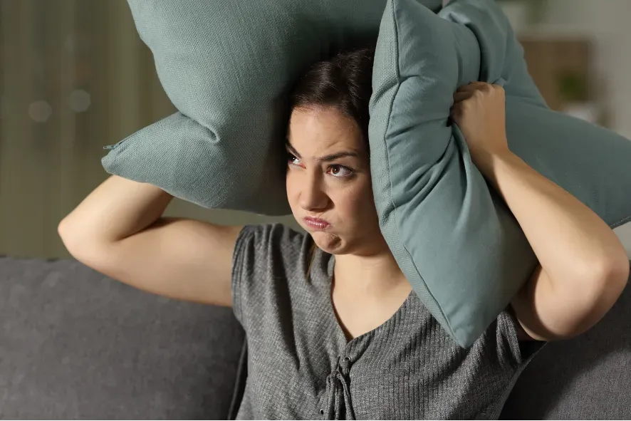 Woman sitting on a couch with pillows pressed over her ears, looking frustrated.