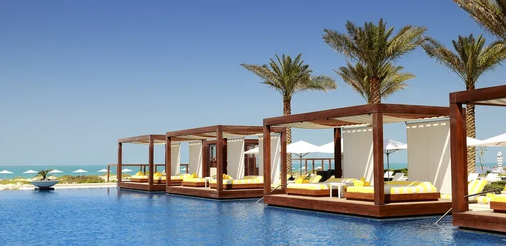 Luxury resort infinity pool with wooden cabanas, palm trees, and ocean view on a sunny beach