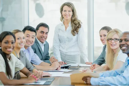 Business team seated around a conference table with a woman standing at the head in a bright meeting room