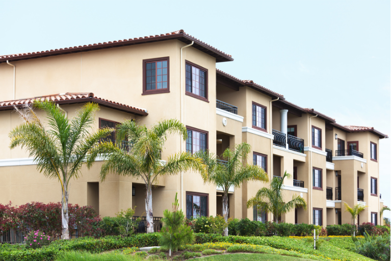 Tan stucco apartment building with palm trees and green landscaping under a bright sky