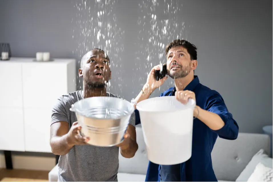 Two men catch water in buckets during an indoor leak, looking up in a living room