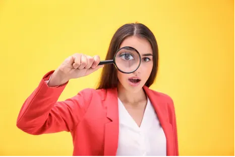Woman in pink blazer holding a magnifying glass over one eye against a yellow background