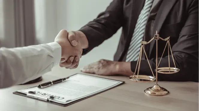 Handshake between two people at a desk with a legal contract clipboard and scales of justice.