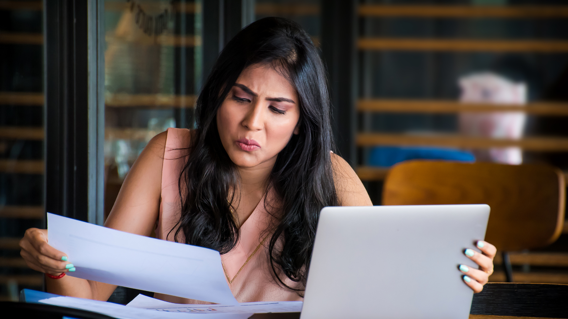 Woman reviewing papers beside a laptop at a table, looking concerned in a cafe or office setting