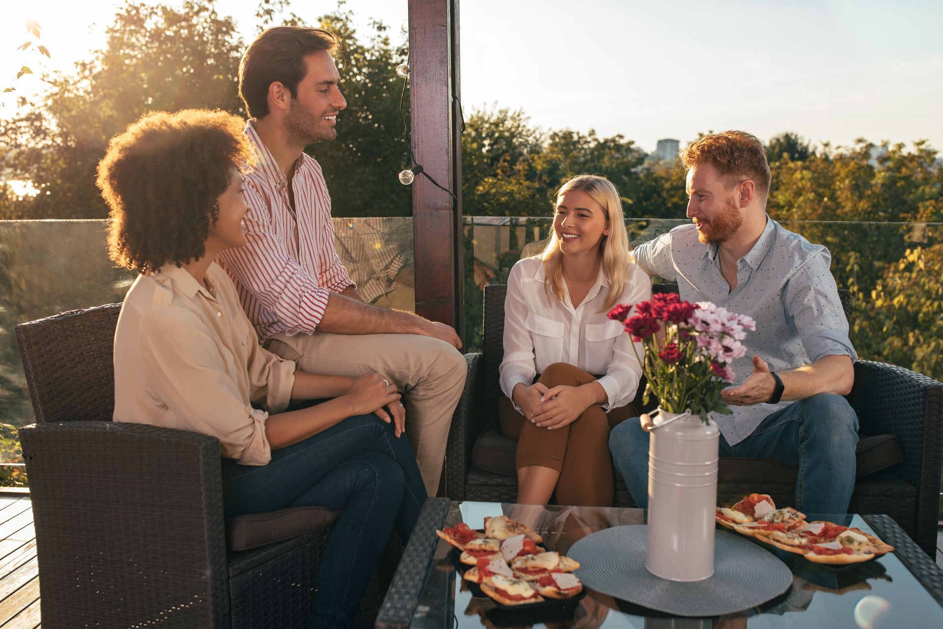 Four friends chatting on a rooftop patio at sunset with drinks, flowers, and appetizers on a table