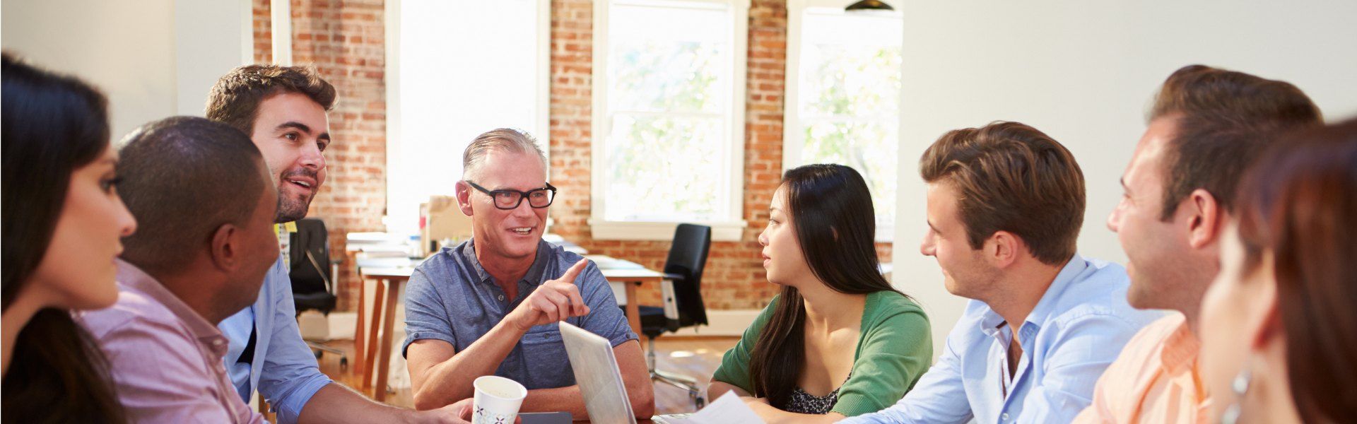 People in a meeting around a table, talking and taking notes in a bright office.