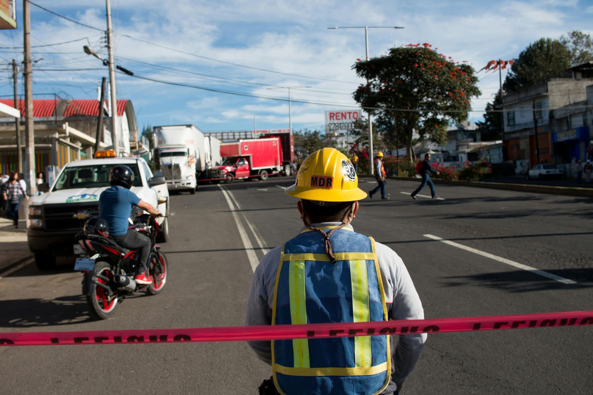 Worker in yellow hard hat and blue vest directing traffic on a road near trucks and motorcycles.