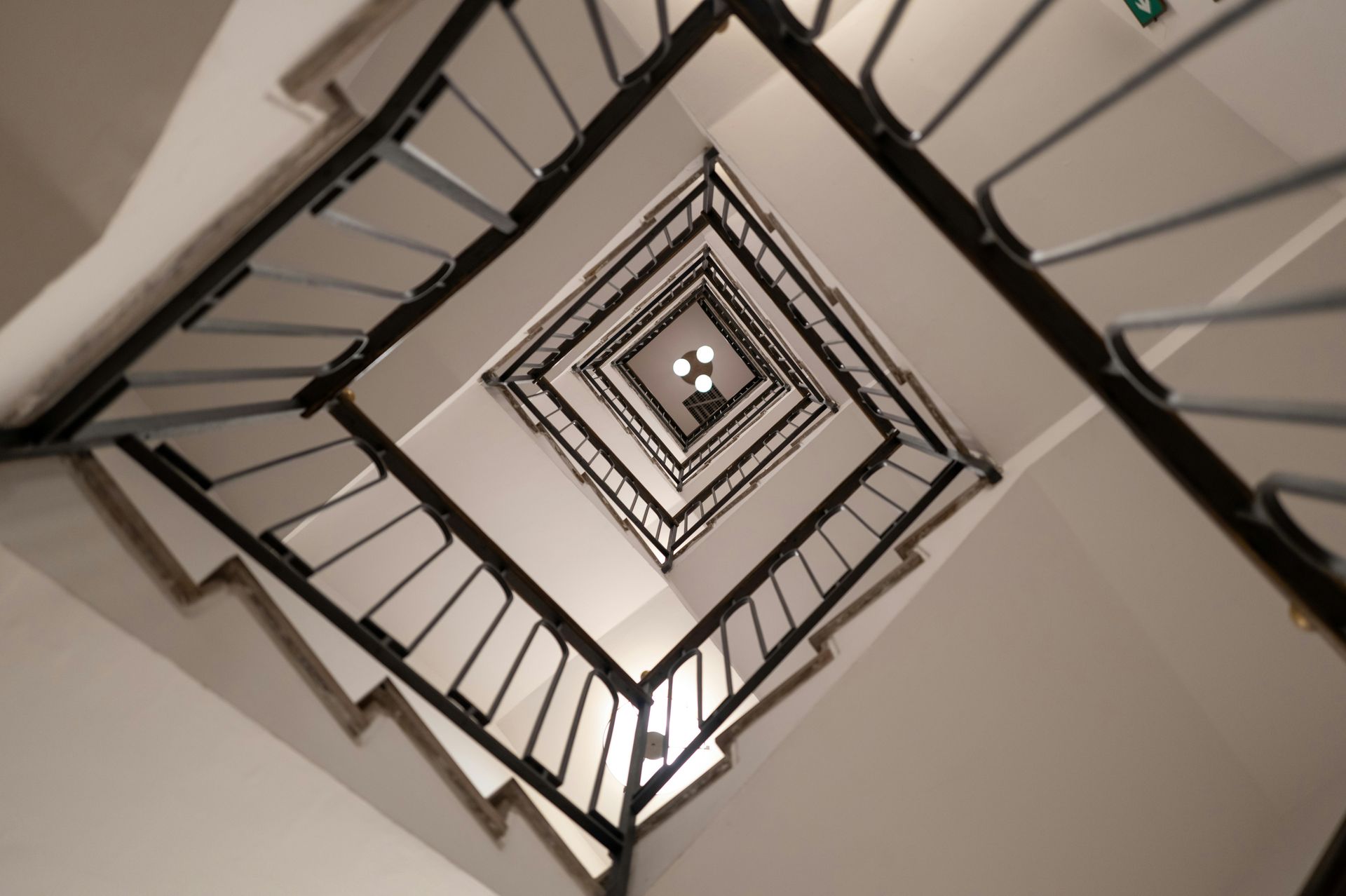 Spiral view up a square stairwell with black railings and white walls, converging to a bright center.