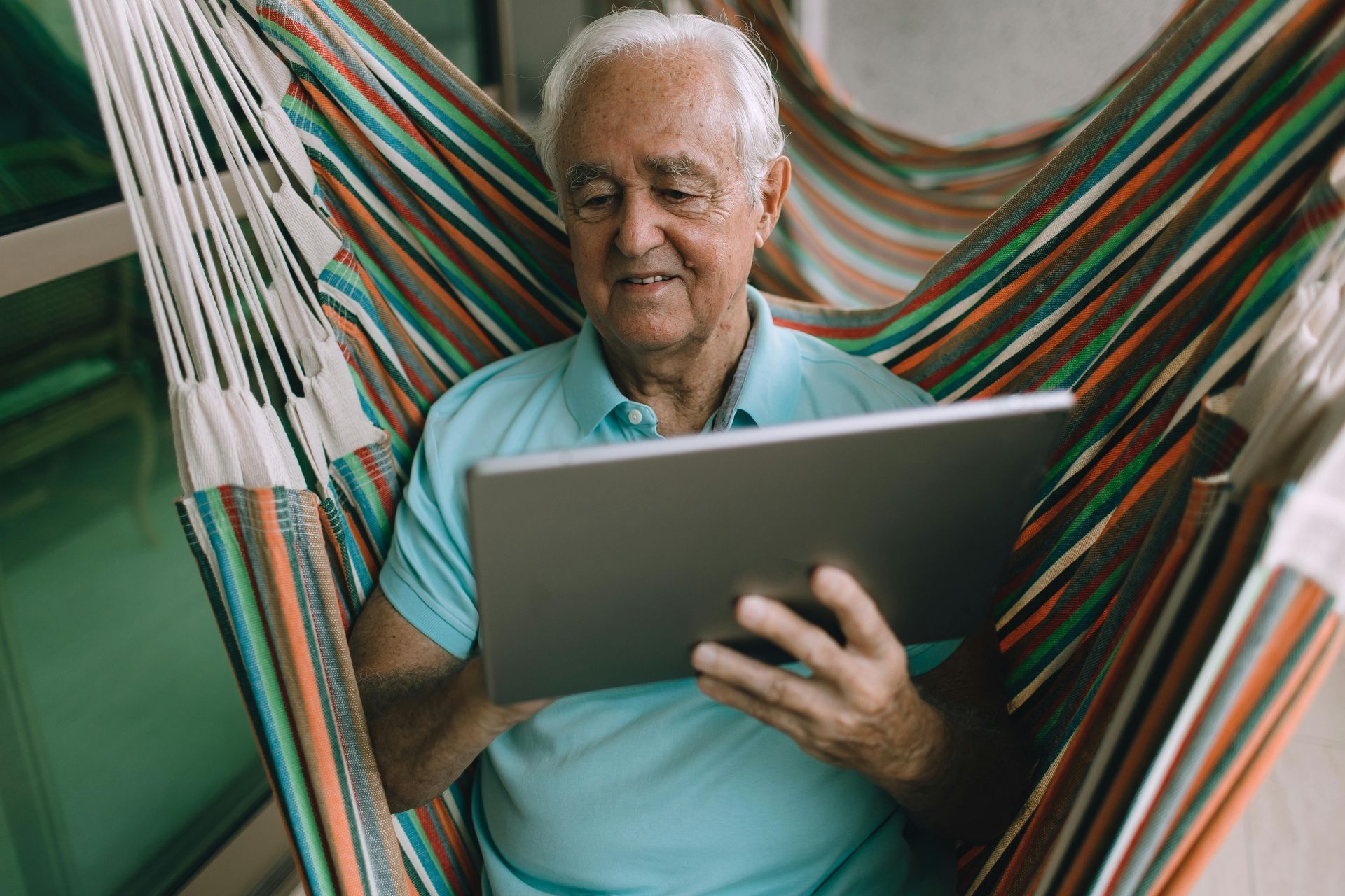 Man holding a laptop in a colorful hammock, wearing a light blue shirt and looking focused