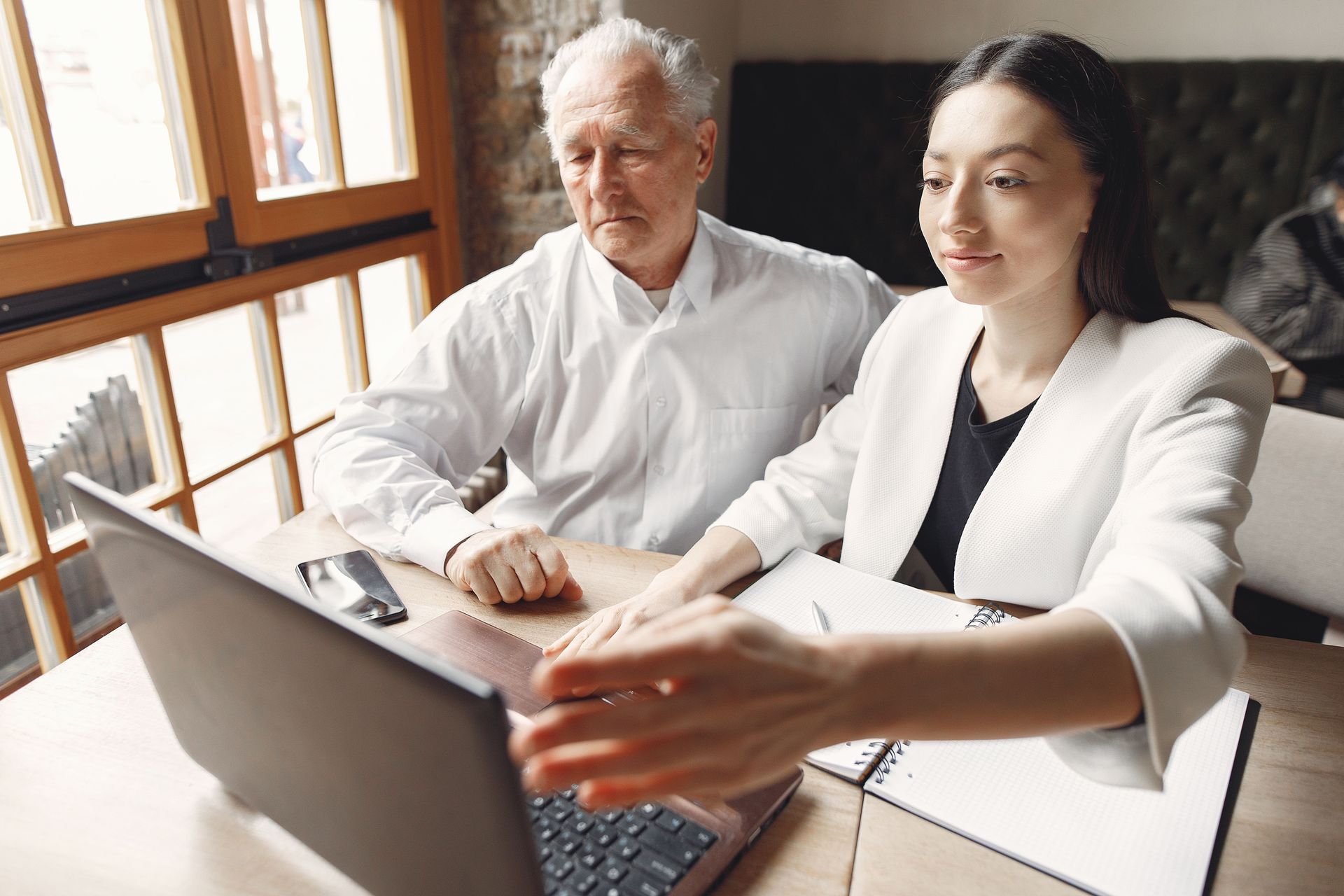 Two people reviewing a laptop at a table near a window, with a notebook in front of them.