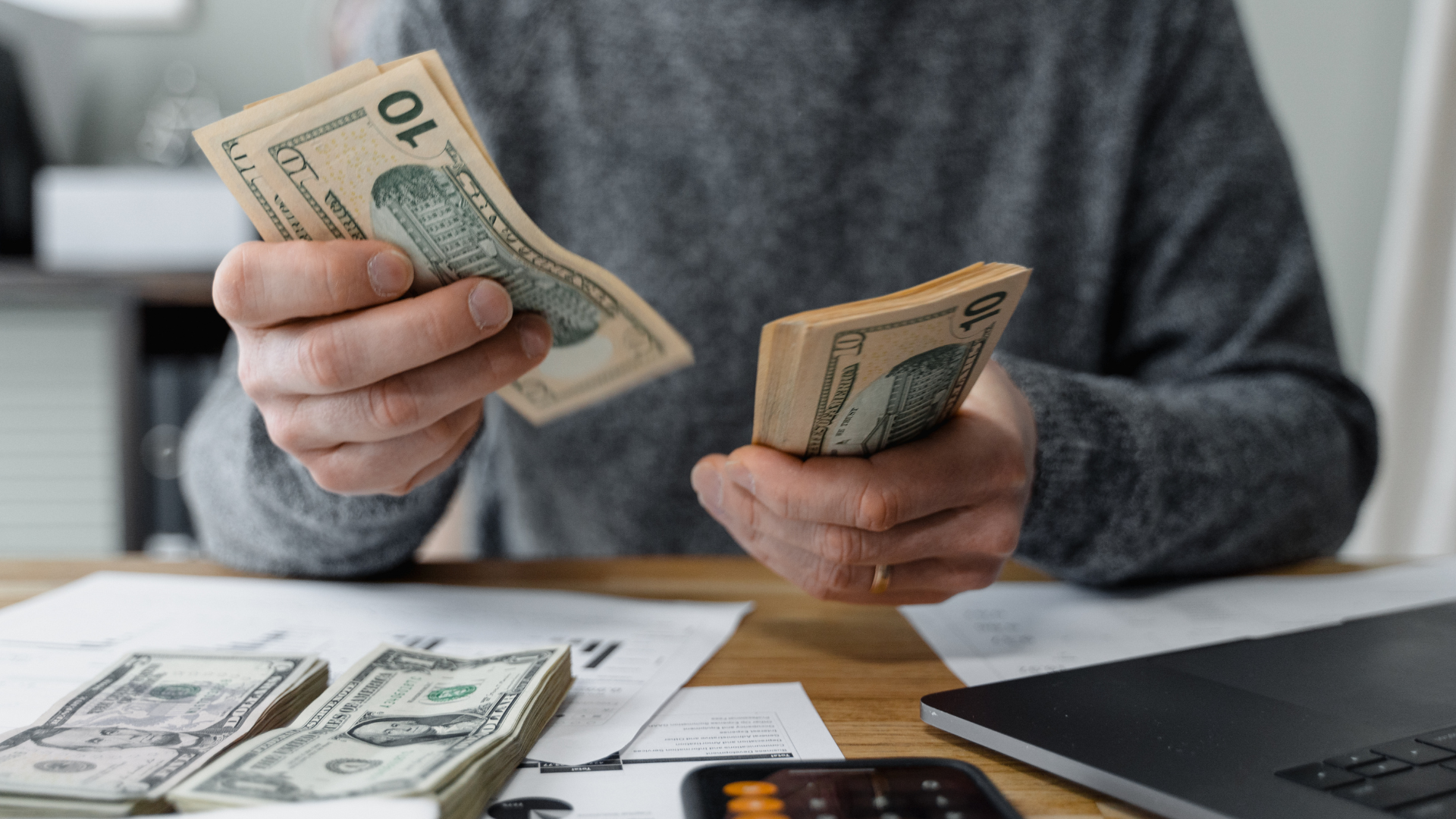 Person counting dollar bills over a desk with paperwork and a calculator