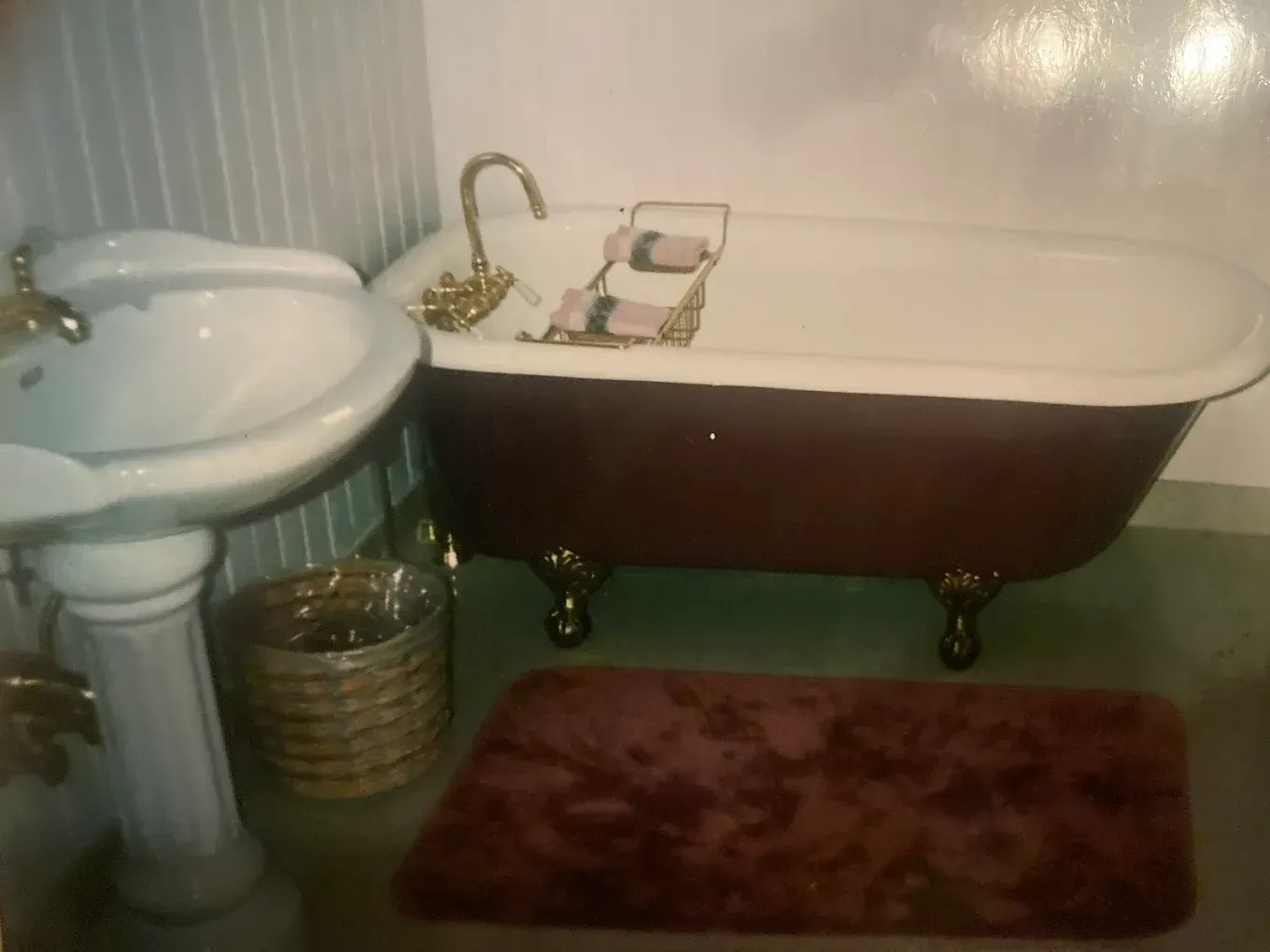 Small vintage bathroom with white pedestal sink, clawfoot tub, brass fixtures, and burgundy bath mat.