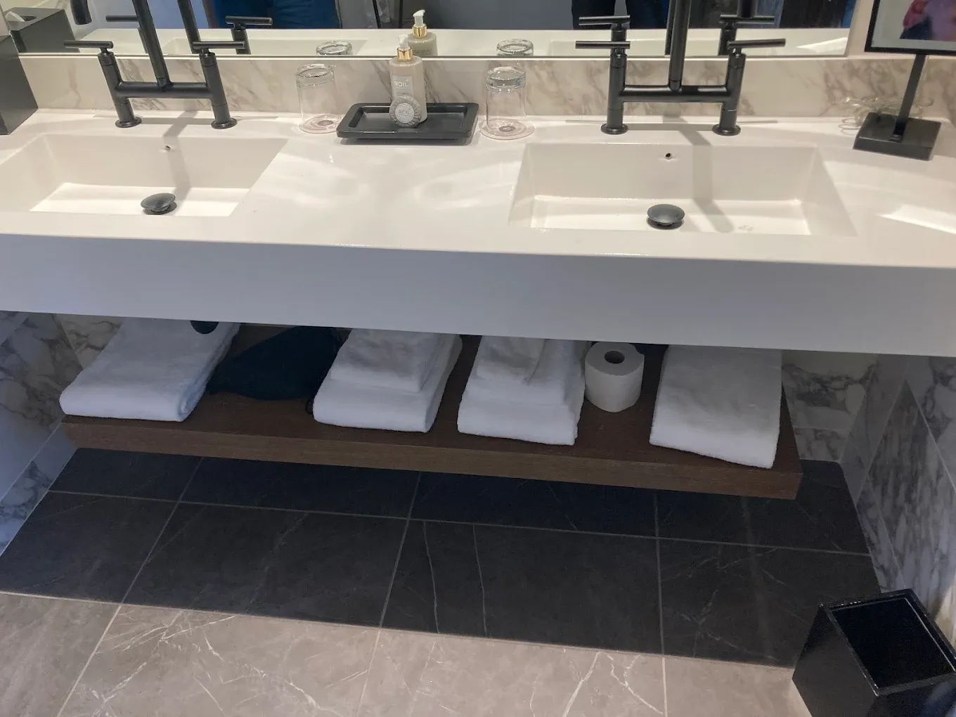Bathroom sink counter with four faucets, folded white towels on a shelf below, and dark tile flooring.