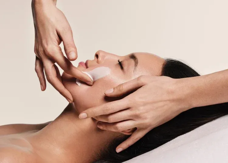 Woman receiving facial treatment, hands applying cream to cheek, eyes closed, light background.