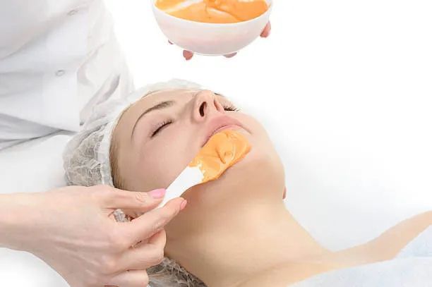 Person receiving a facial treatment; an esthetician applies an orange mask to the face.