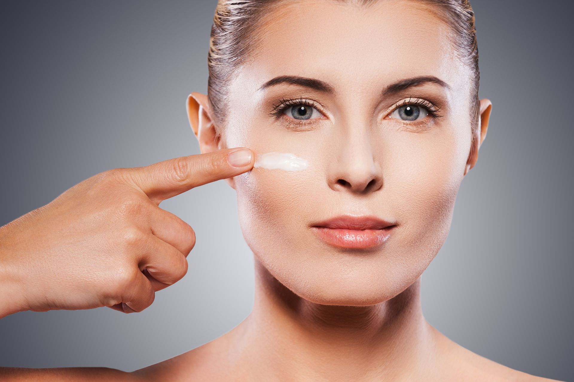 Woman applying cream to her cheek with a finger, close-up, gray background.