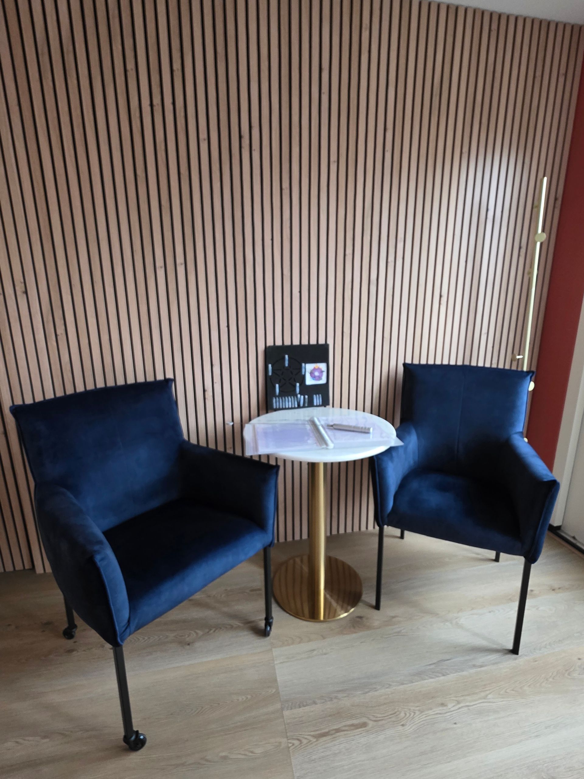 Twee blauwe fluwelen stoelen staan ​​tegenover een kleine witte tafel met documenten, tegen een wand van houten latten.