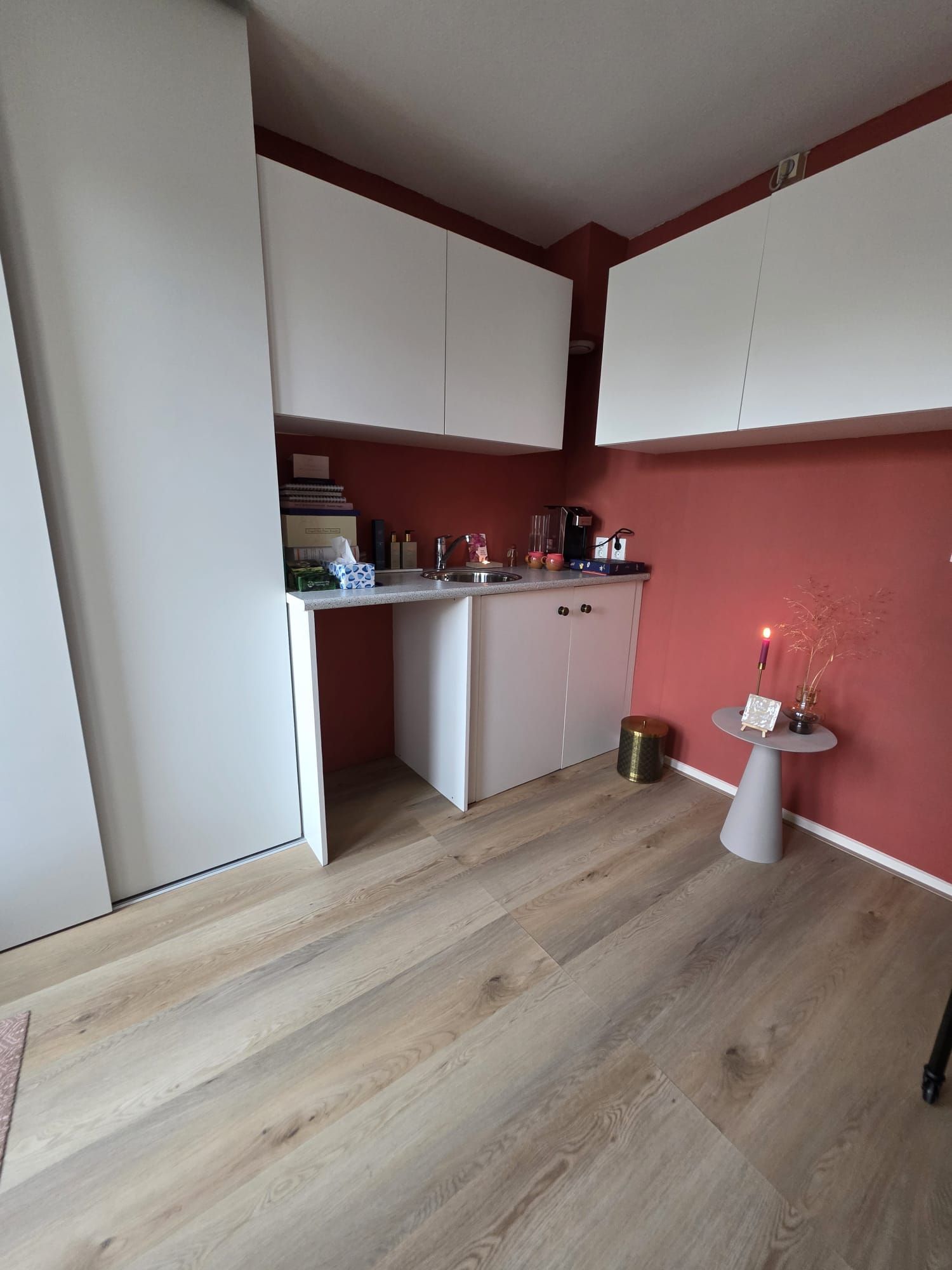 Een kleine, witte keuken met rode muren en een houten vloer. Keukenkastjes boven een aanrechtblad met apparatuur en decoratie.